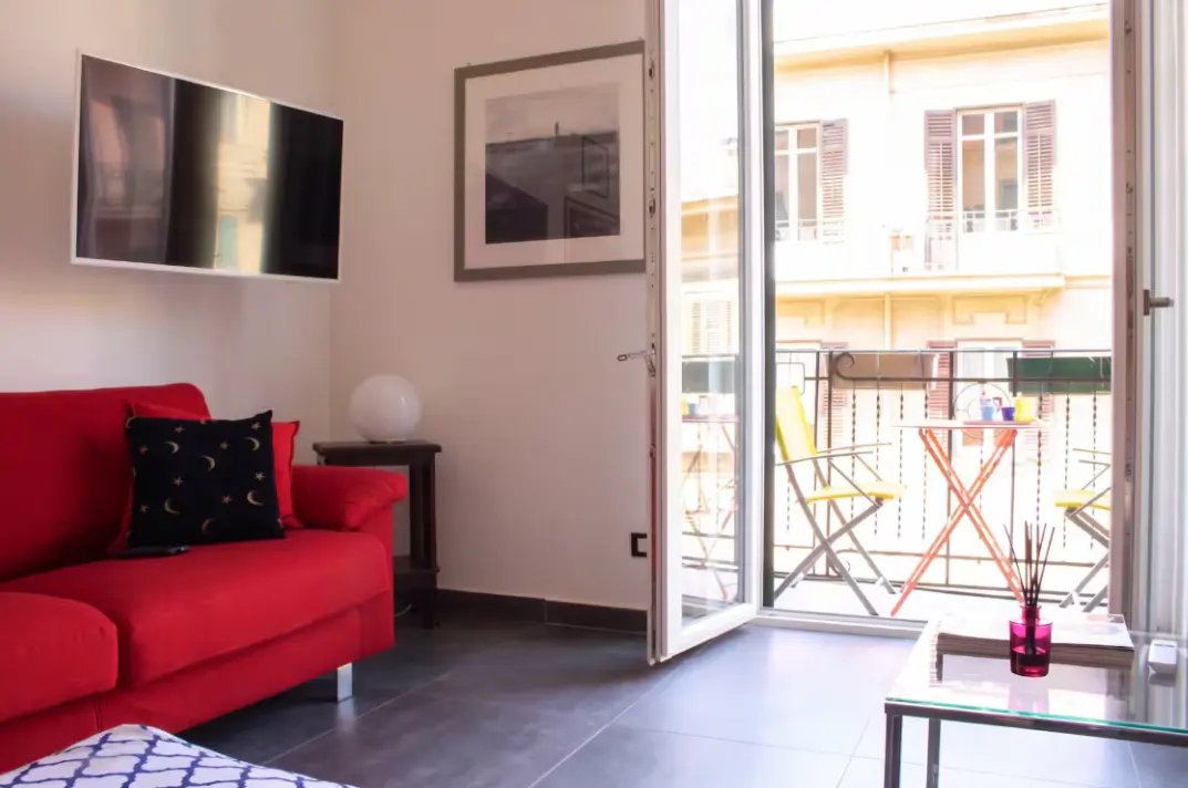 Bright living room with a red sofa, TV, and open balcony. Balcony has a table and chairs with a view of a building across the street.