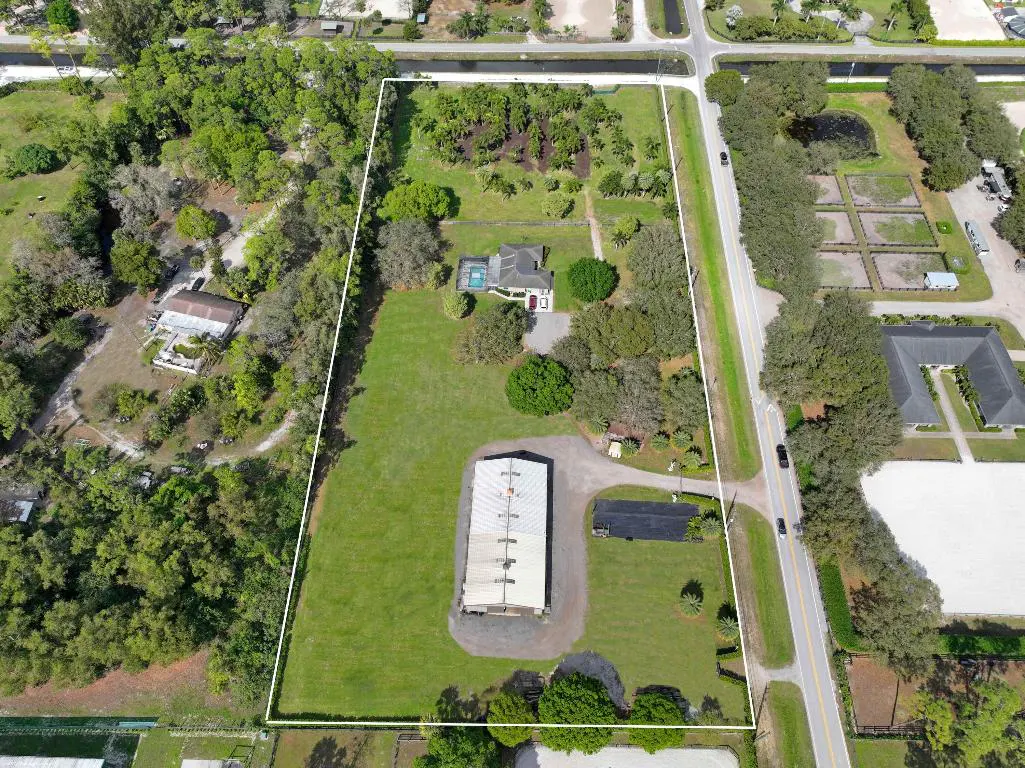 Aerial view of a large property with a house, pool, barn, and green lawn, bordered by trees and a road.