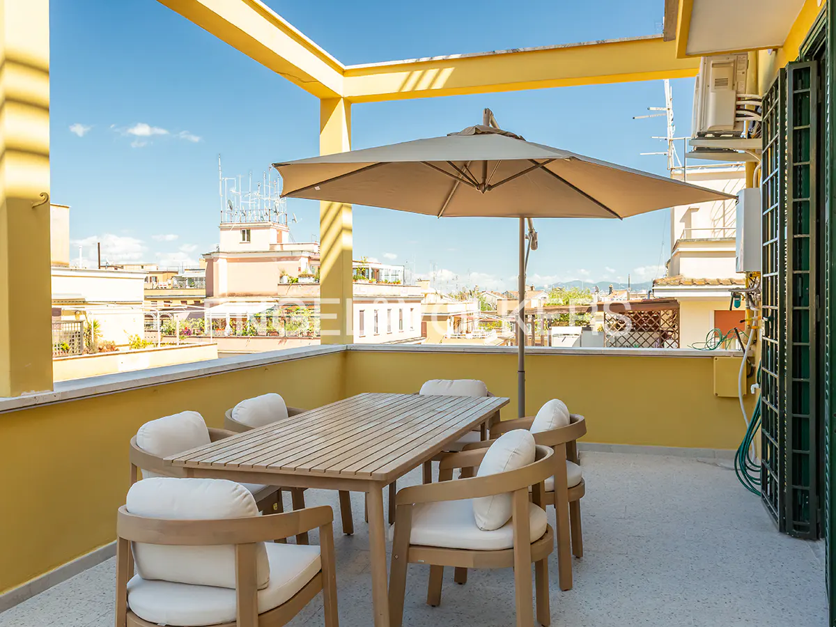Outdoor patio with a wooden table, six chairs, and a beige umbrella. Yellow walls and a city view in the background.