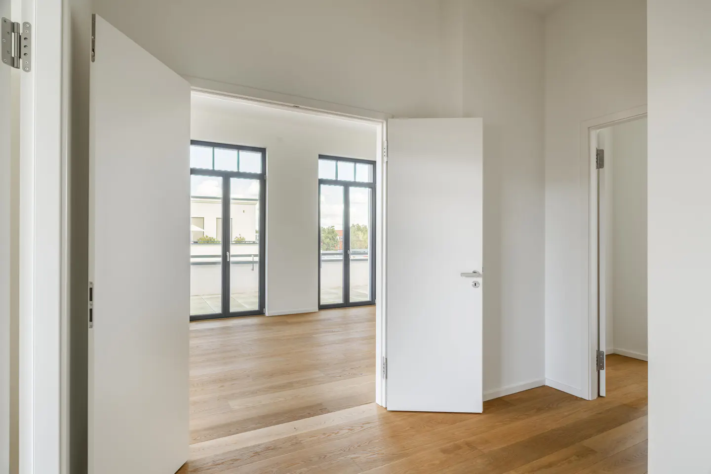 A bright, modern room with hardwood floors and white walls. Two sets of black-framed glass doors lead to a balcony. White doors are ajar.