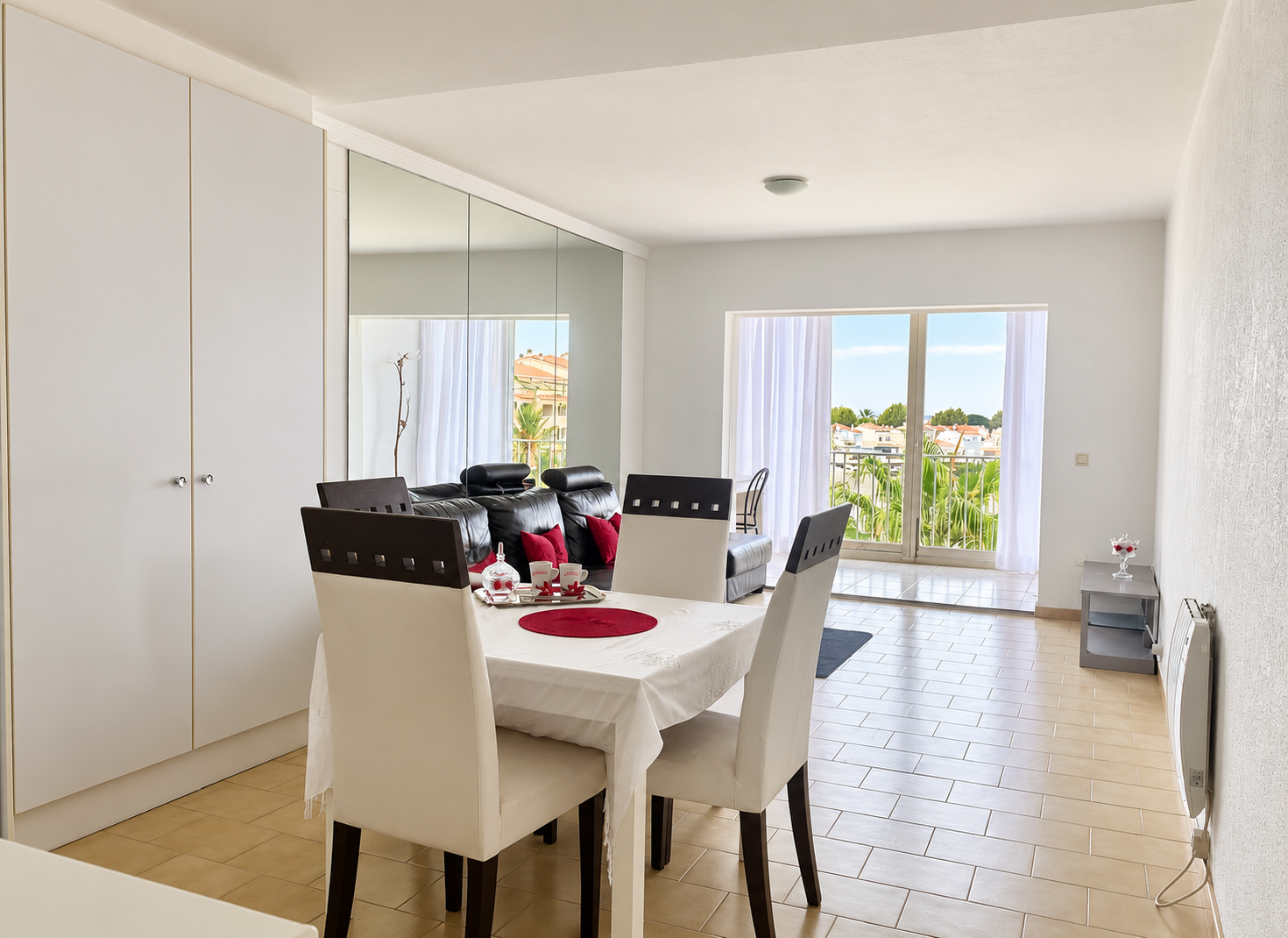 Bright dining area with a white table, four chairs, mirrored wall, and a balcony view of palm trees and buildings. [AI-generated text]