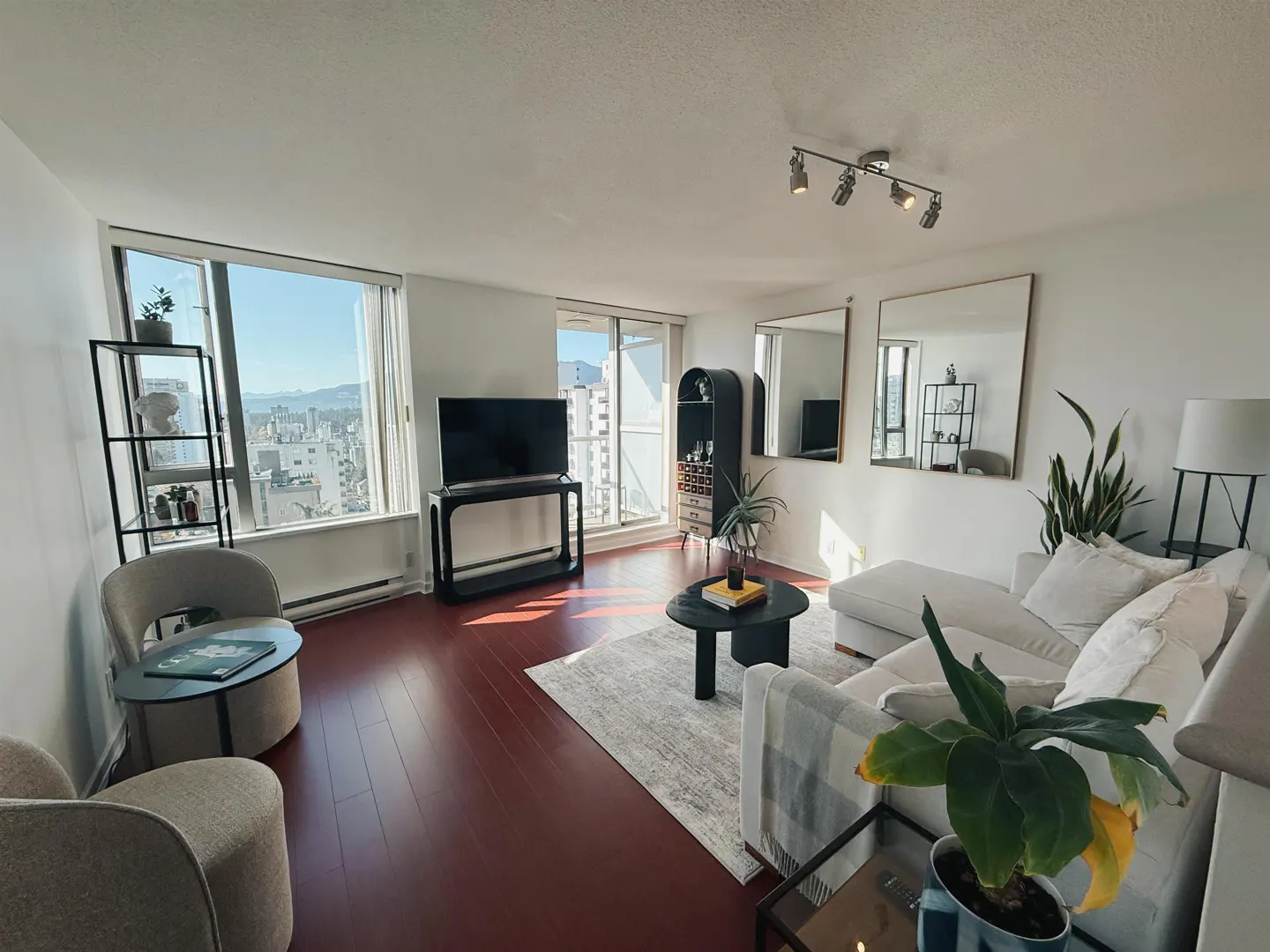 Bright living room with white walls, red wood floors, and a large window with a city view. A white sofa, black TV stand, and plants add modern touches.