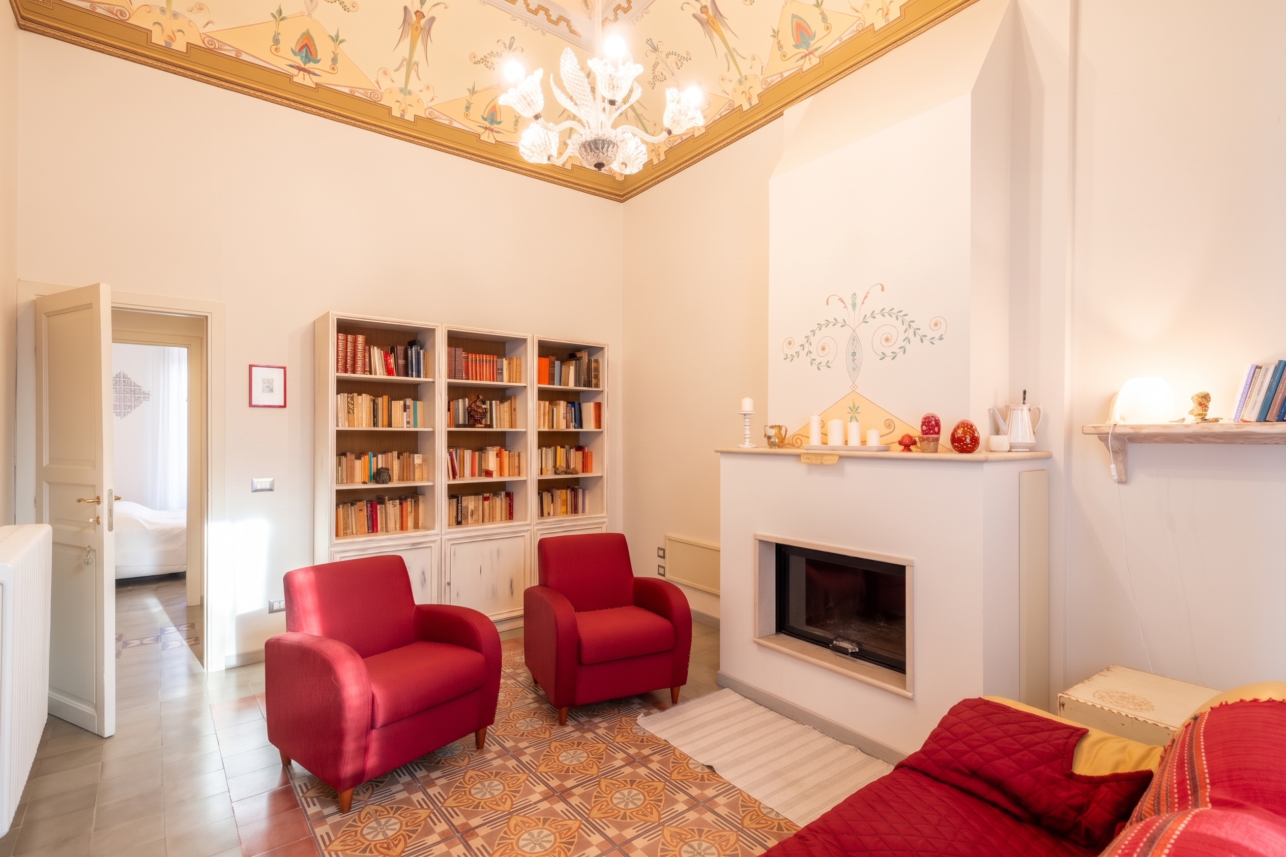Living room with two red armchairs, a fireplace, a large bookcase, and a decorative painted ceiling with a chandelier. [AI-generated text]