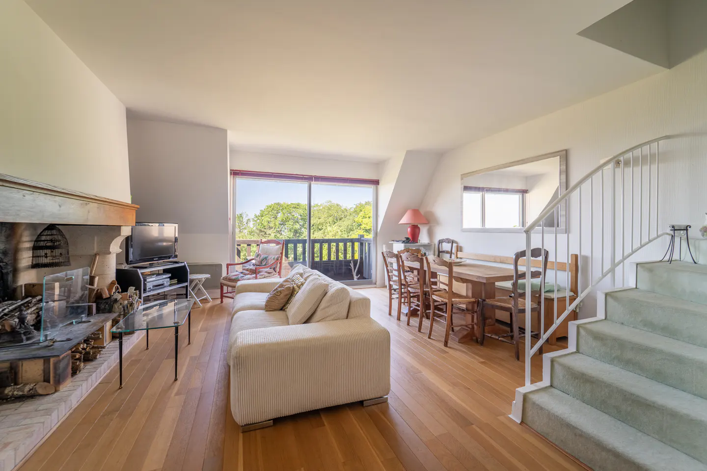 Bright living room with hardwood floors, a large fireplace, a cream sofa, a dining table, and a staircase. Balcony with green trees visible.