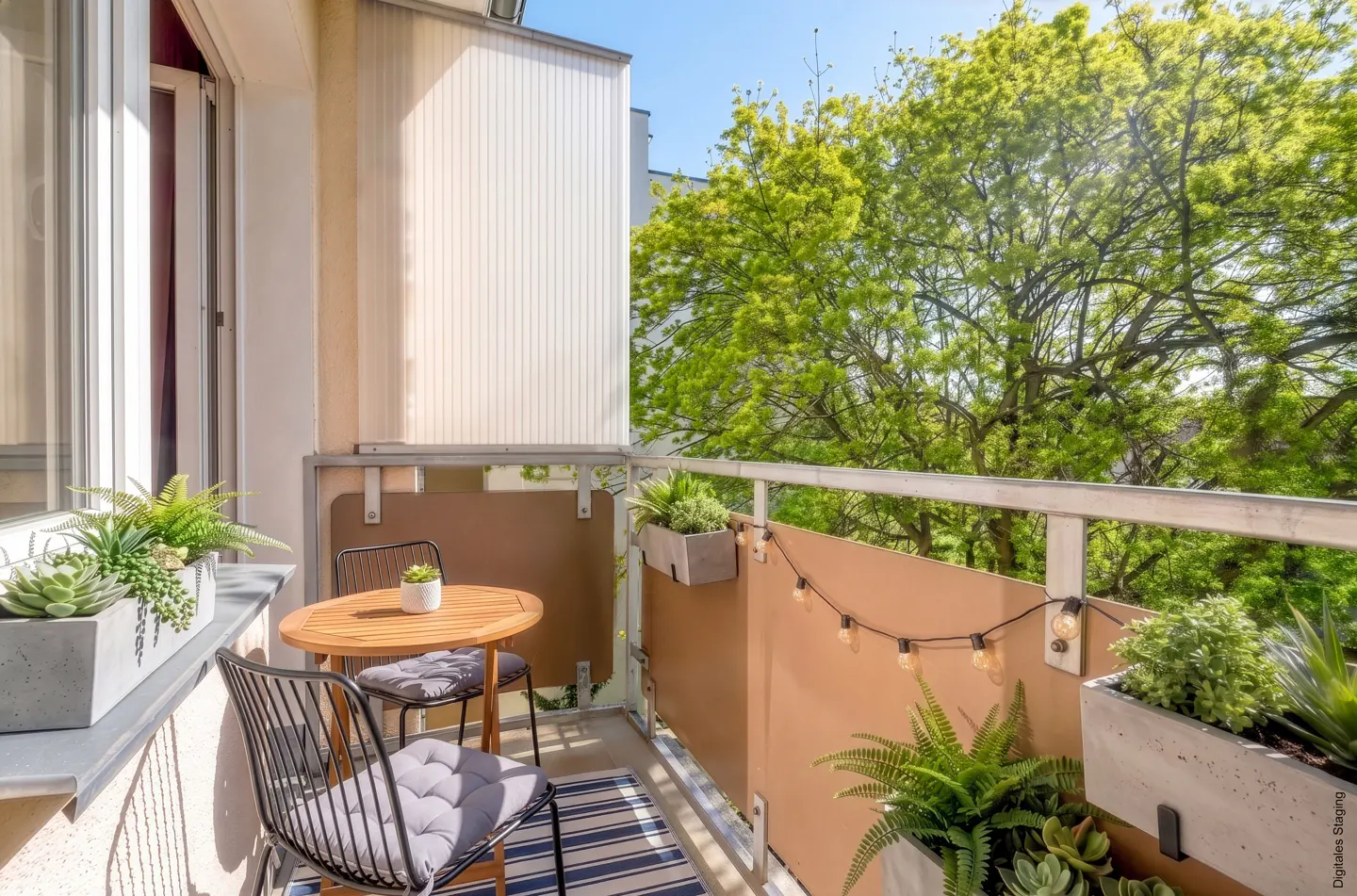 Balcony with a round wooden table, two black chairs with gray cushions, and potted plants. String lights hang along the railing.