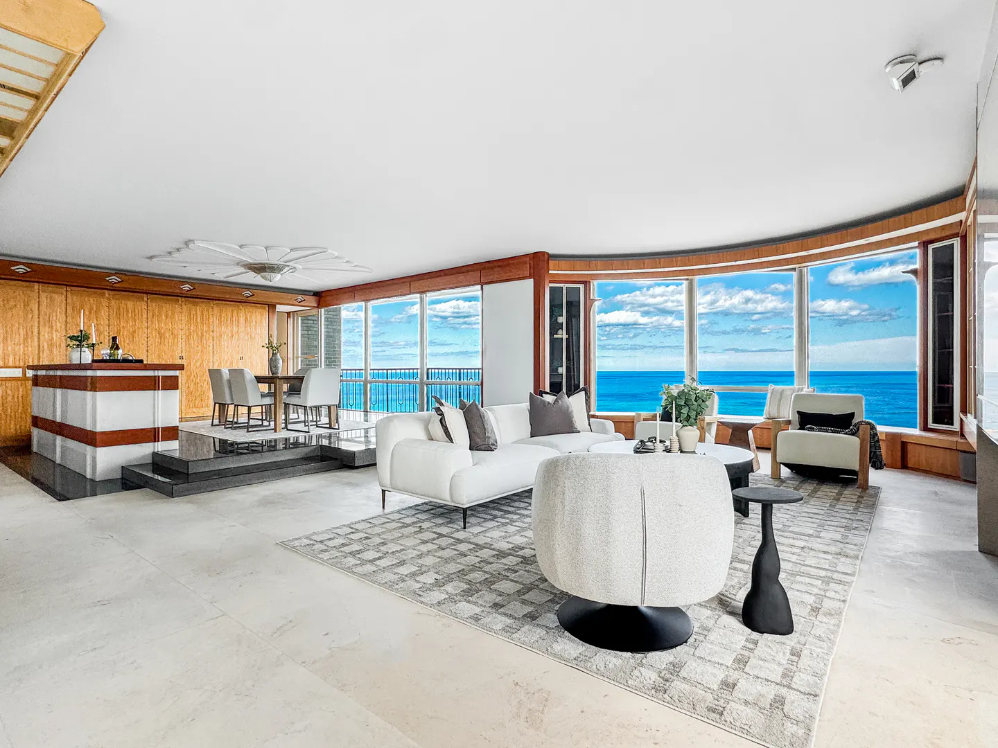 Bright, open living room with ocean view. White sofa and chairs sit on a patterned rug. Bar and dining area in the background.
