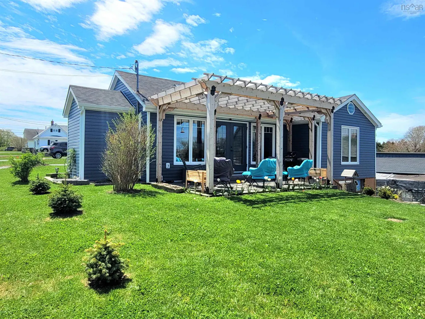 A blue house with a pergola over a patio with blue chairs, on a green lawn under a blue sky.