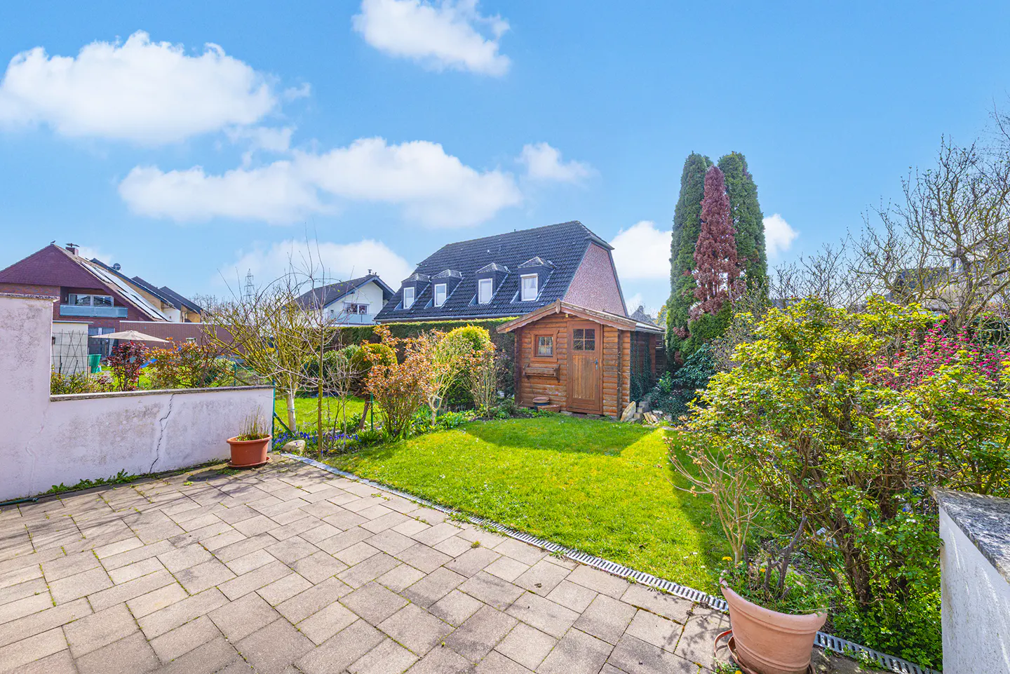 A backyard with a stone patio, green lawn, wooden shed, and a house with dormer windows under a blue sky.