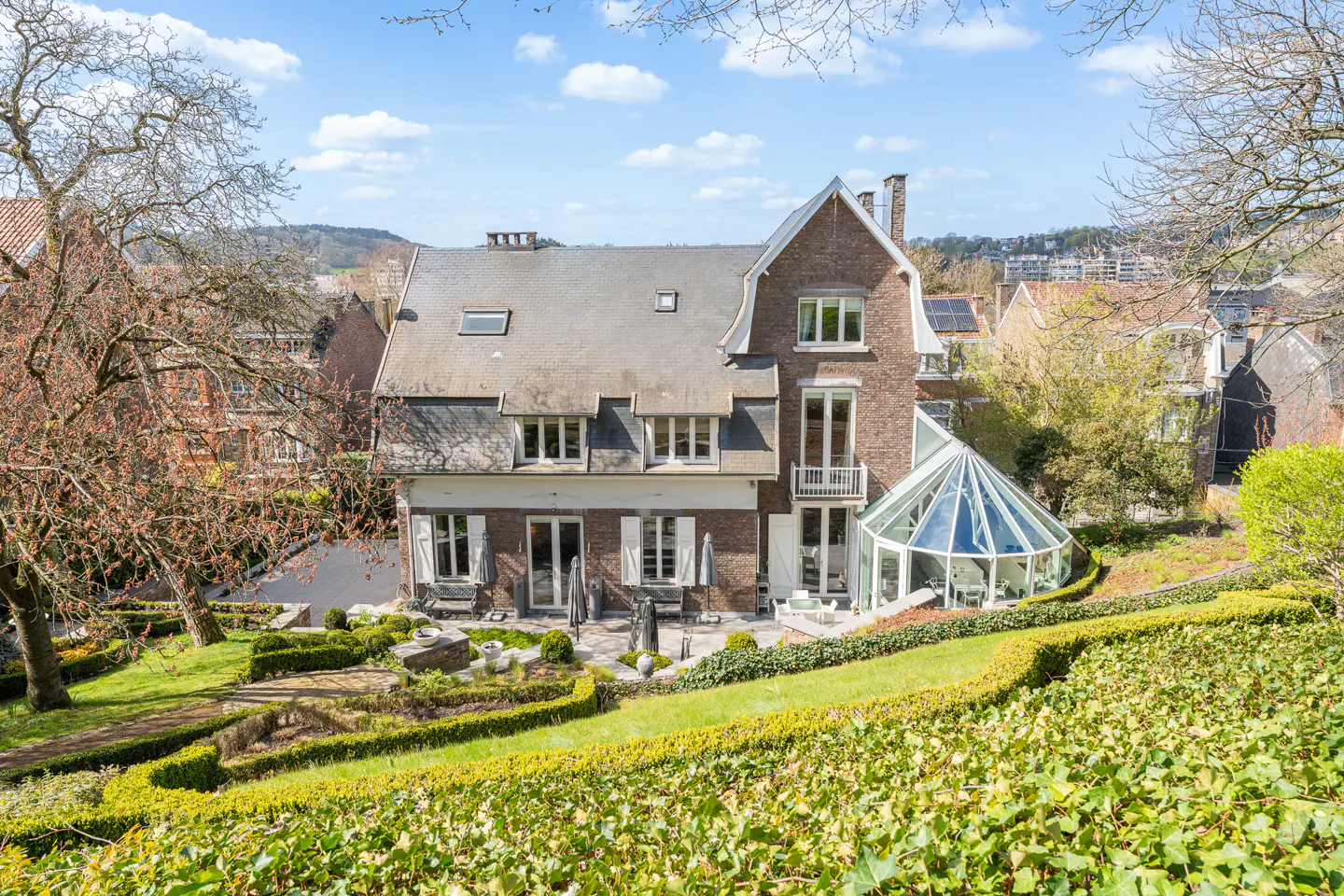 Exterior view of a large brick house with a glass conservatory, a patio, and a tiered garden on a sunny day.