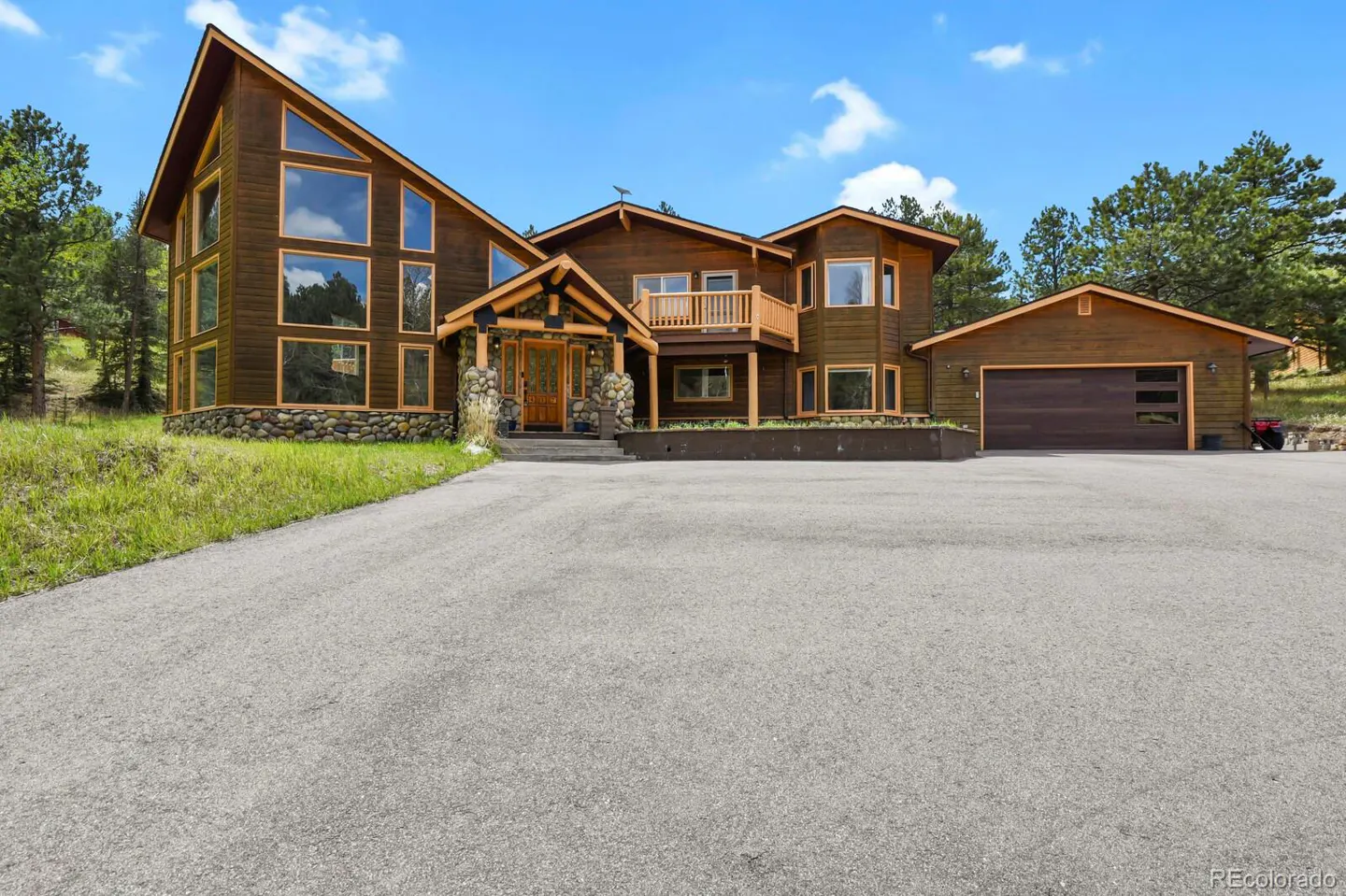 Exterior view of a large, brown, multi-story house with a stone foundation and a gray driveway. Blue sky and trees in the background.
