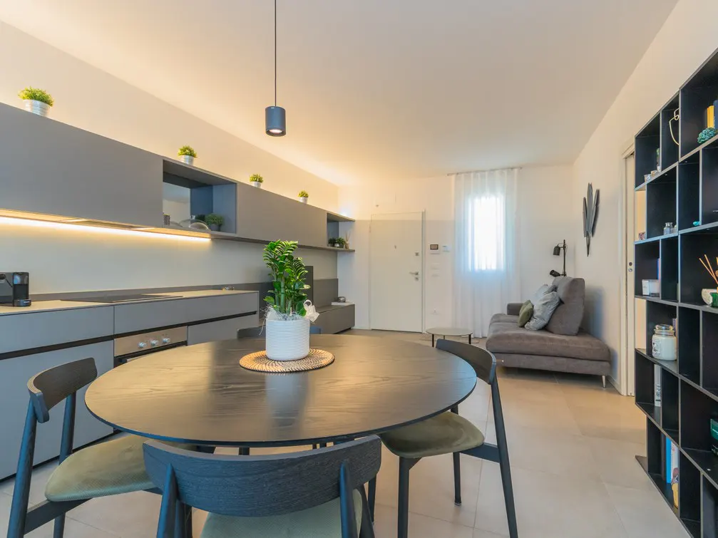 Modern open-plan living area with a round black dining table, grey kitchen cabinets, and a cozy sofa in the background.