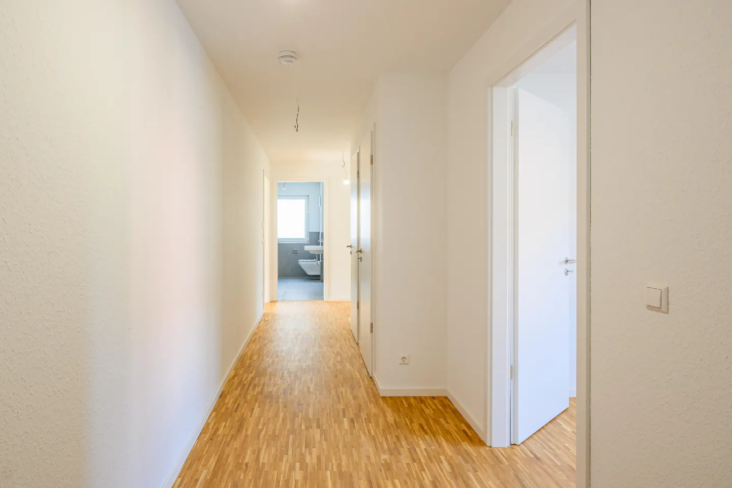 Bright hallway with wood floors and white walls. An open door reveals a bathroom with a toilet and sink.
