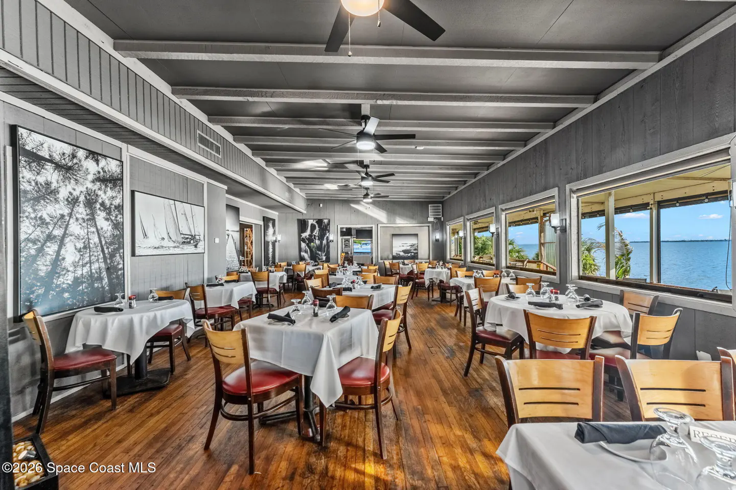 Restaurant dining room with tables set with white linens, wood floors, and water views.