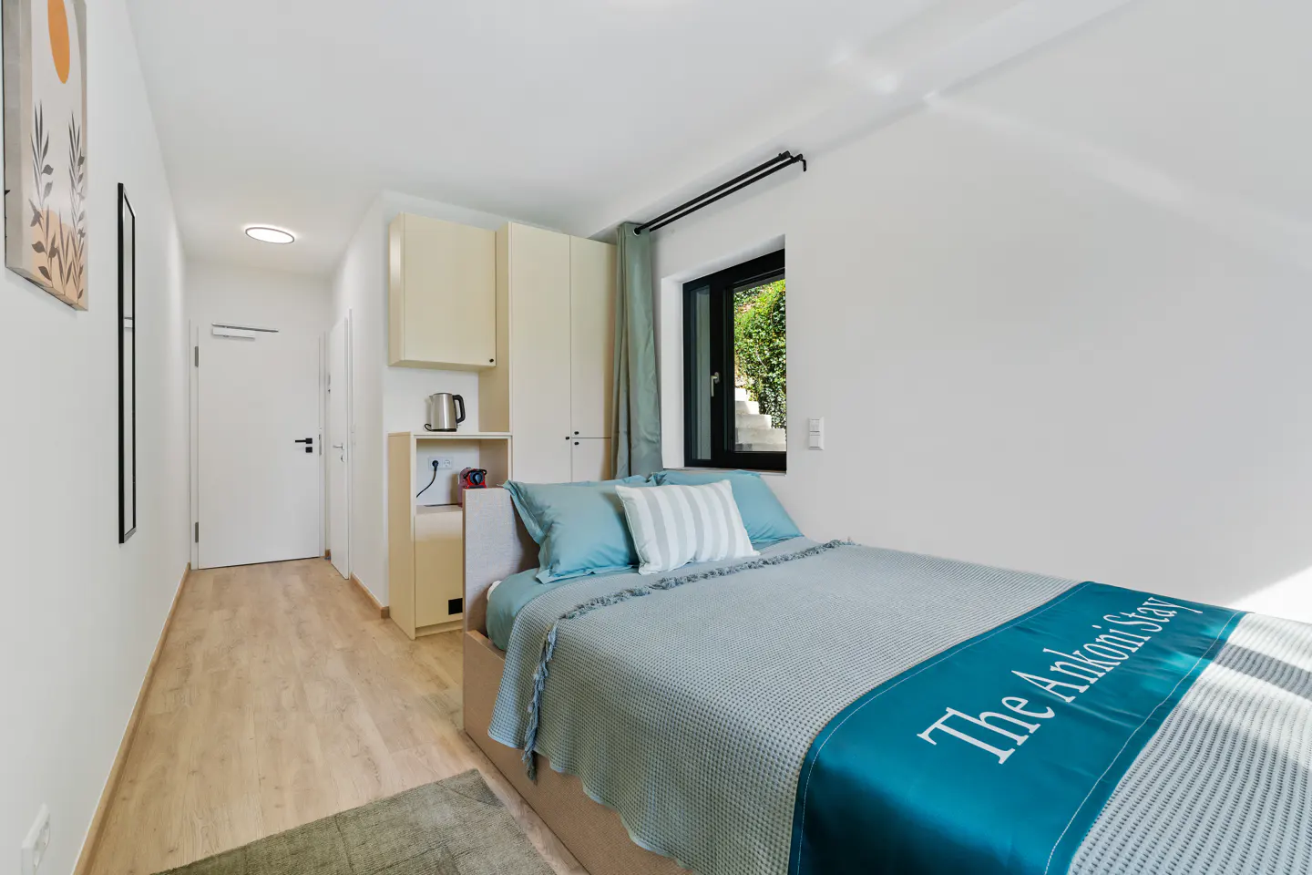 Bright bedroom with a bed, blue pillows, and a teal blanket with "The Ankoni Stay" written on it. There is a window and a small kitchenette.