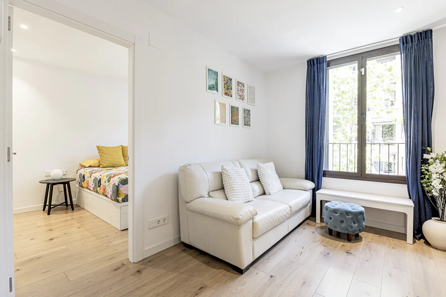 Bright living room with wood floors, white walls, and blue curtains. A white sofa sits near a window with a bench and ottoman. A bedroom is visible through a doorway.
