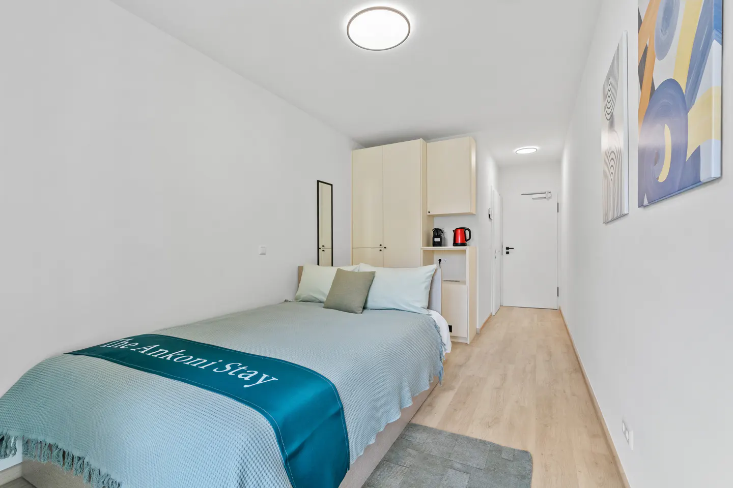 Bright studio apartment with a bed, kitchenette, and white walls. A teal blanket with "The Ankoni Stay" is on the bed. Artwork hangs on the wall.