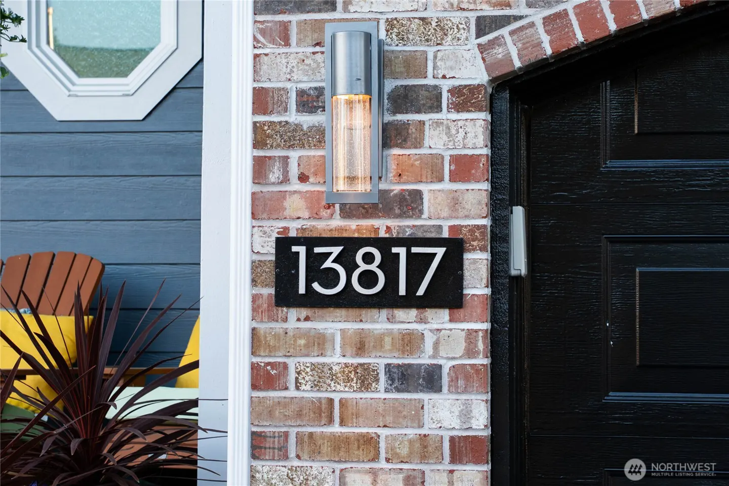 House number 13817 on a brick wall next to a black door, with a modern wall sconce and a glimpse of a patio chair.