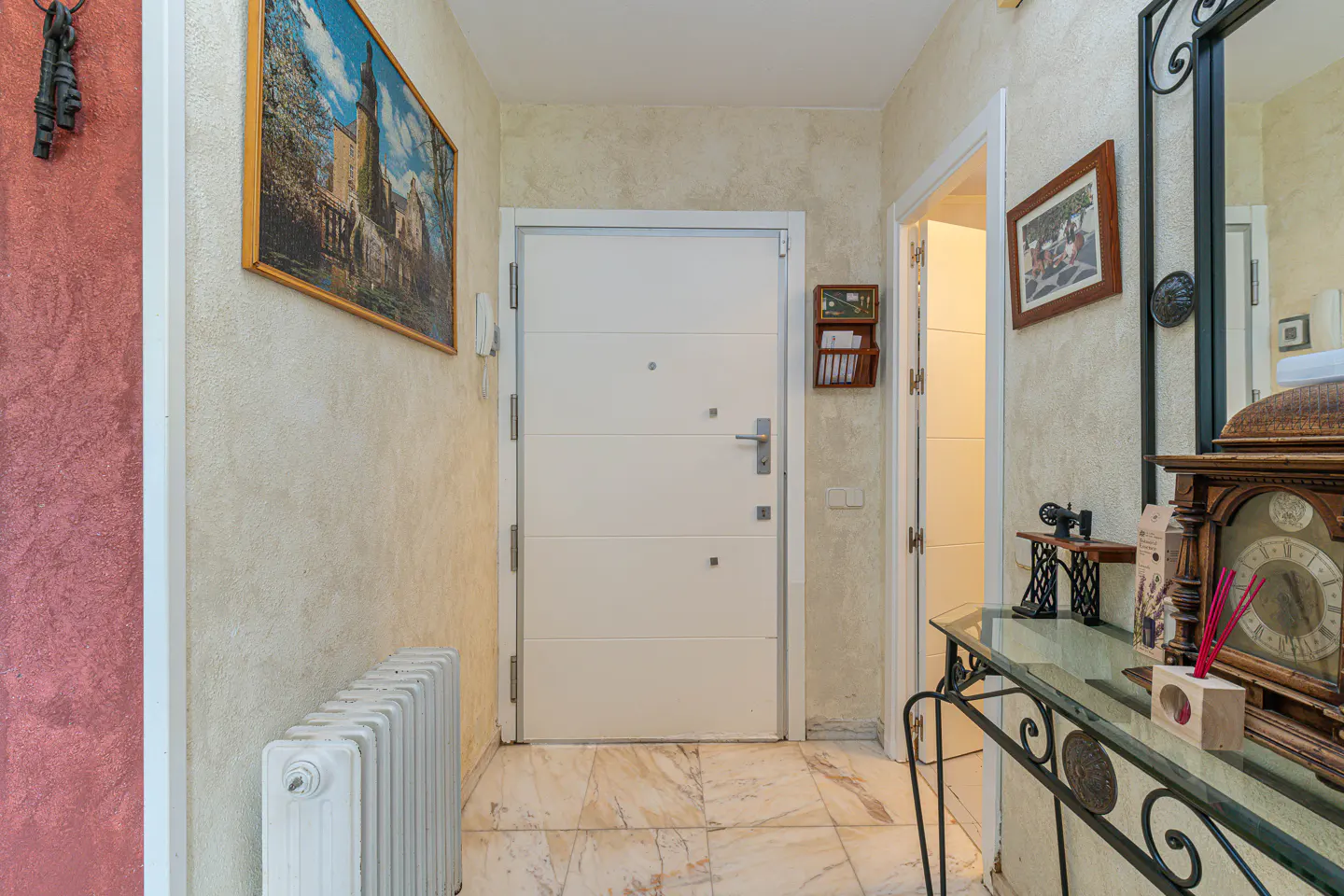 Hallway view with a white front door, marble floor, and textured walls. A radiator sits near a painting of a castle. A console table with a clock and decor is visible.