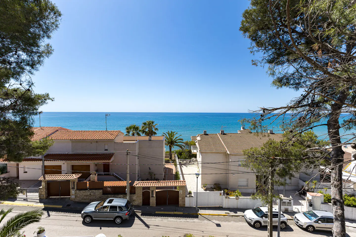 View of a coastal street with beige houses, parked cars, and pine trees overlooking the blue Mediterranean Sea.