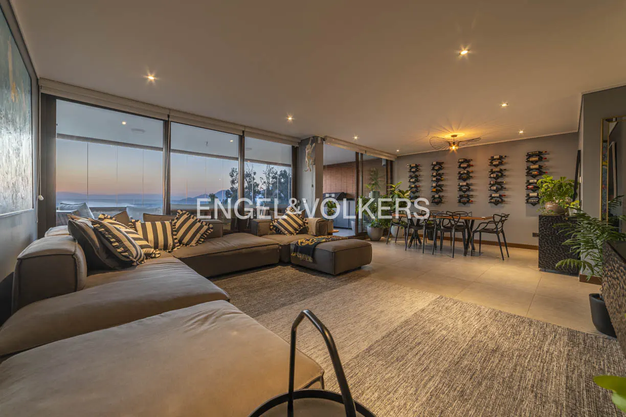 Spacious living room with a large gray sectional sofa, floor-to-ceiling windows with ocean views, and a dining area with wine racks.