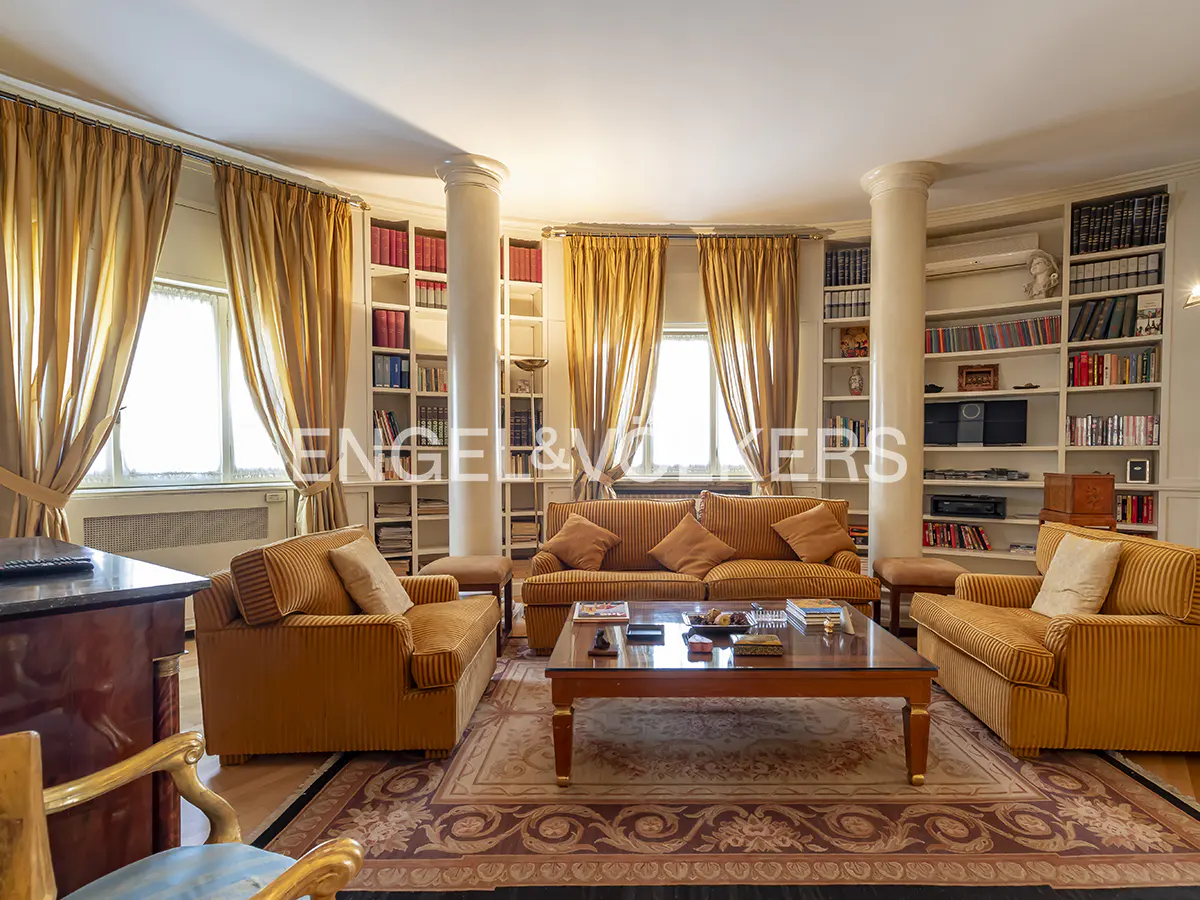 A living room with orange sofas, a coffee table, and bookshelves filled with books. Curtains frame the windows.