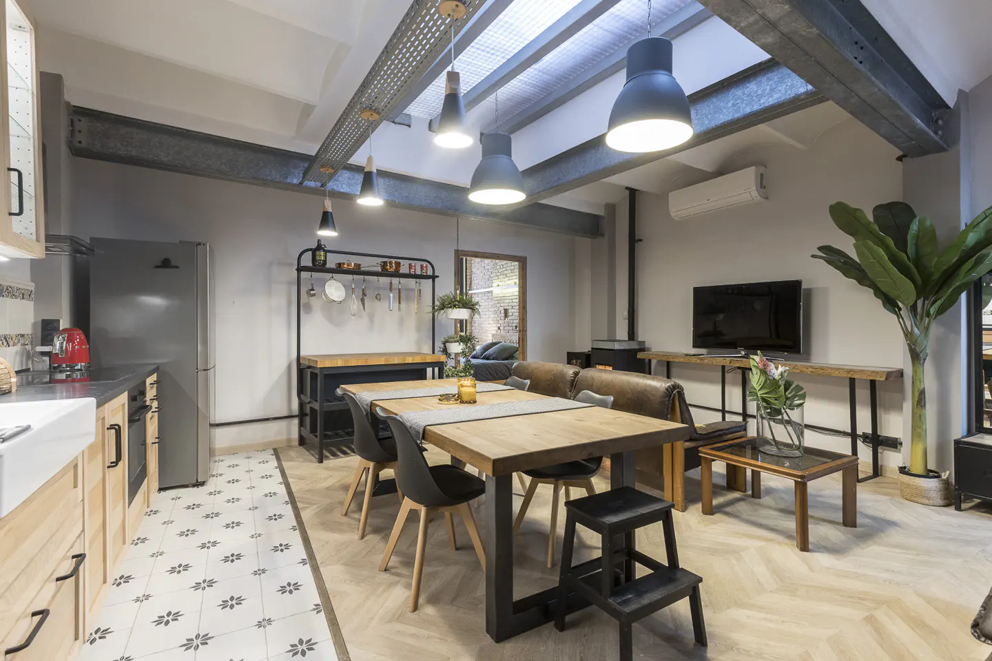 Open-concept living space with kitchen, dining, and living areas. Wood table, black chairs, and industrial lighting. Neutral tones with pops of greenery.