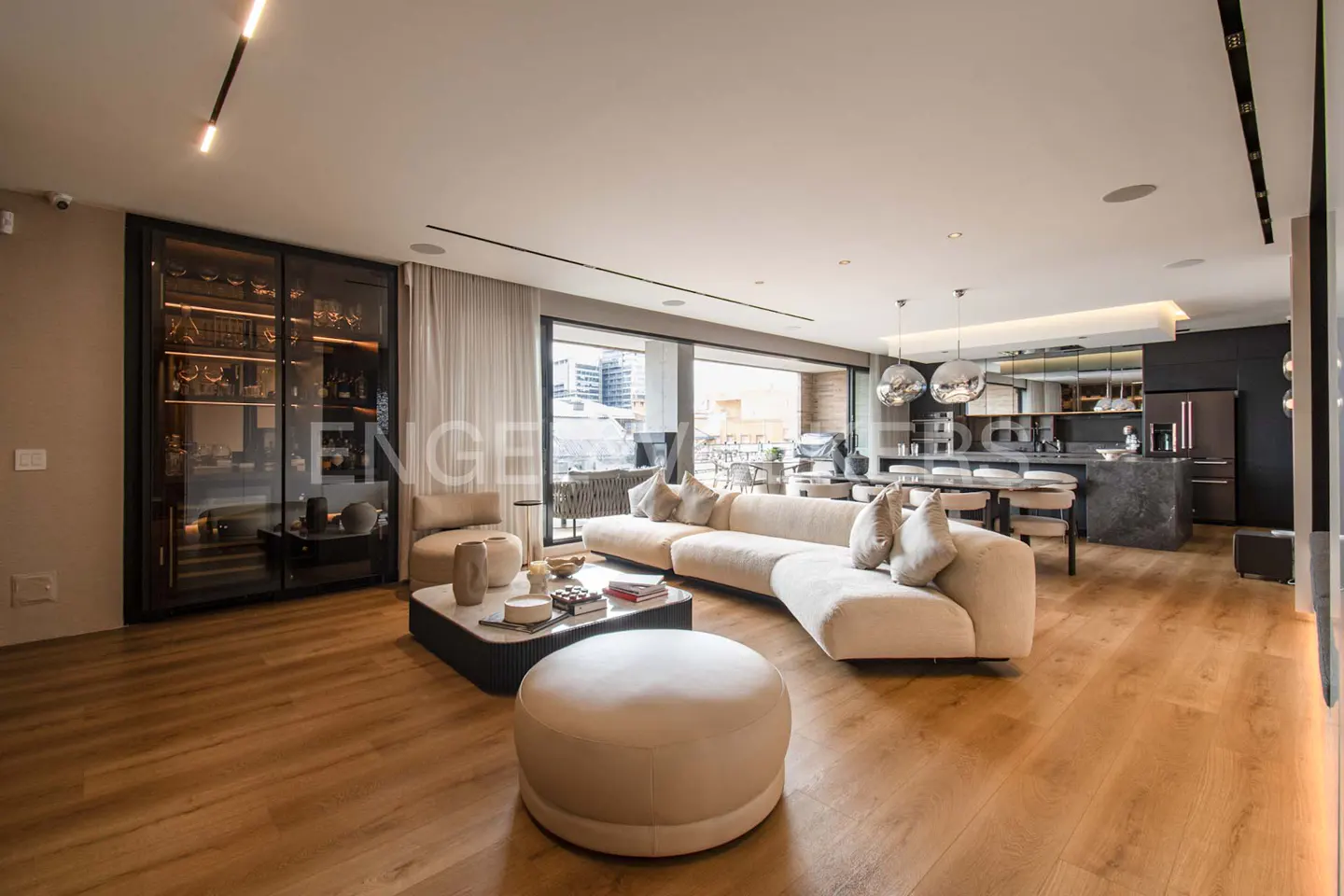 Open-concept living space with wood floors, a cream sectional sofa, and a glass-front wine cabinet. A kitchen is visible in the background.