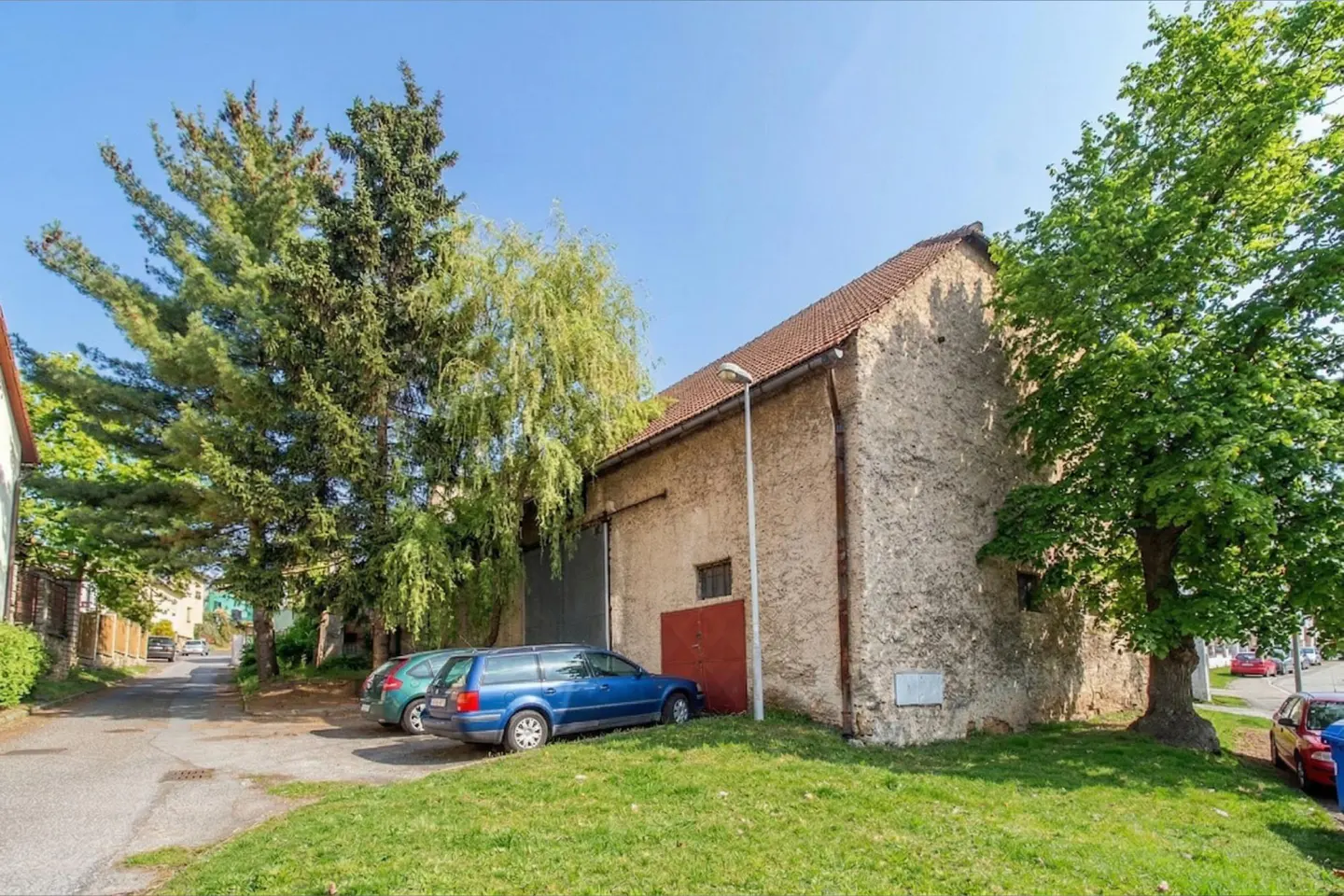 Exterior view of a tan stone building with a red door, a brown roof, and green trees. Two cars are parked in front of the building.