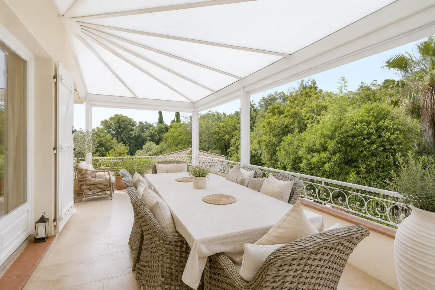 Covered patio with a long table, wicker chairs, and white cushions. Lush green trees are visible in the background.