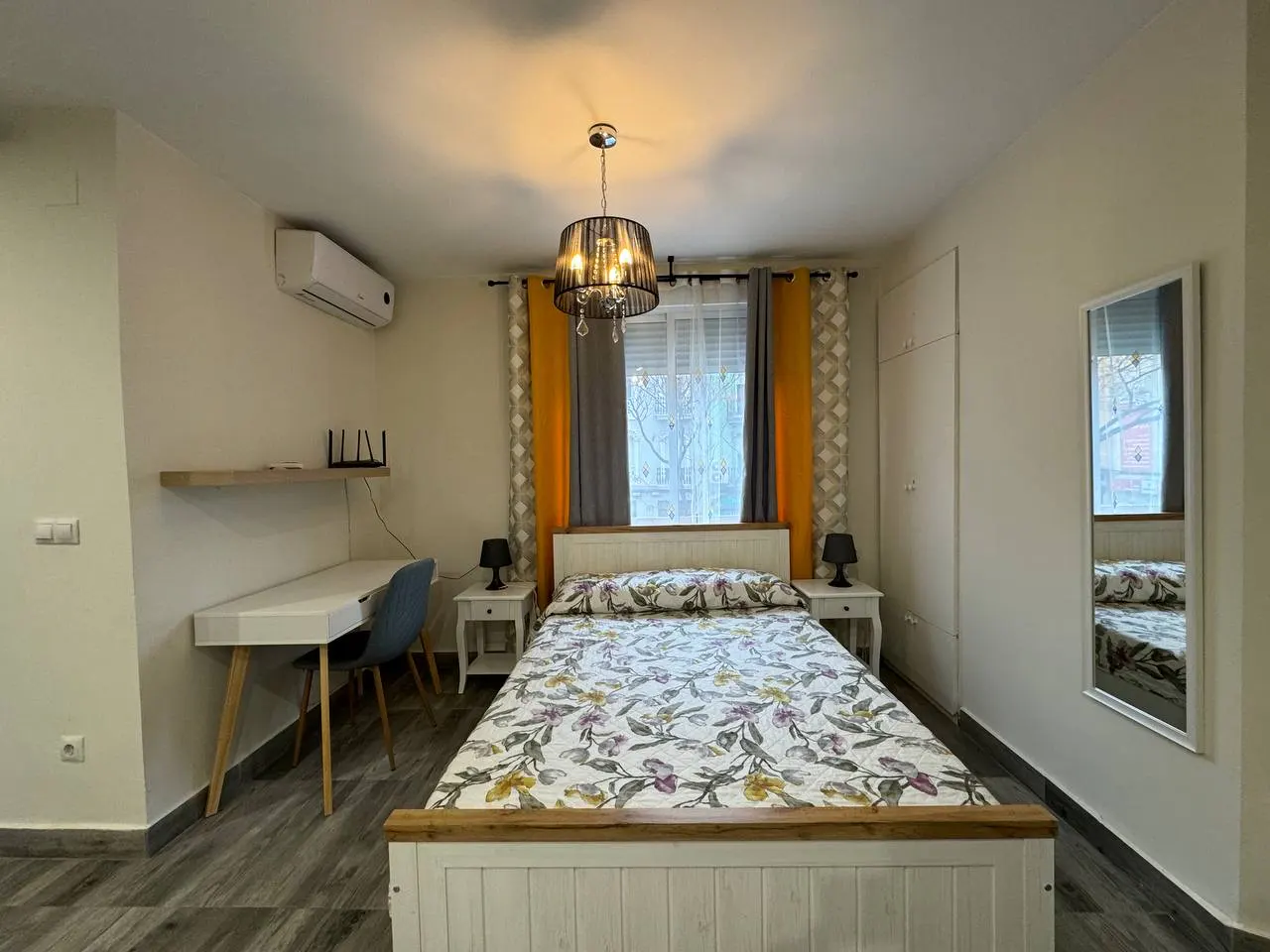 Bedroom with a white bed, floral bedding, desk, chair, and a black chandelier. Curtains are yellow and gray. A mirror reflects the bed.