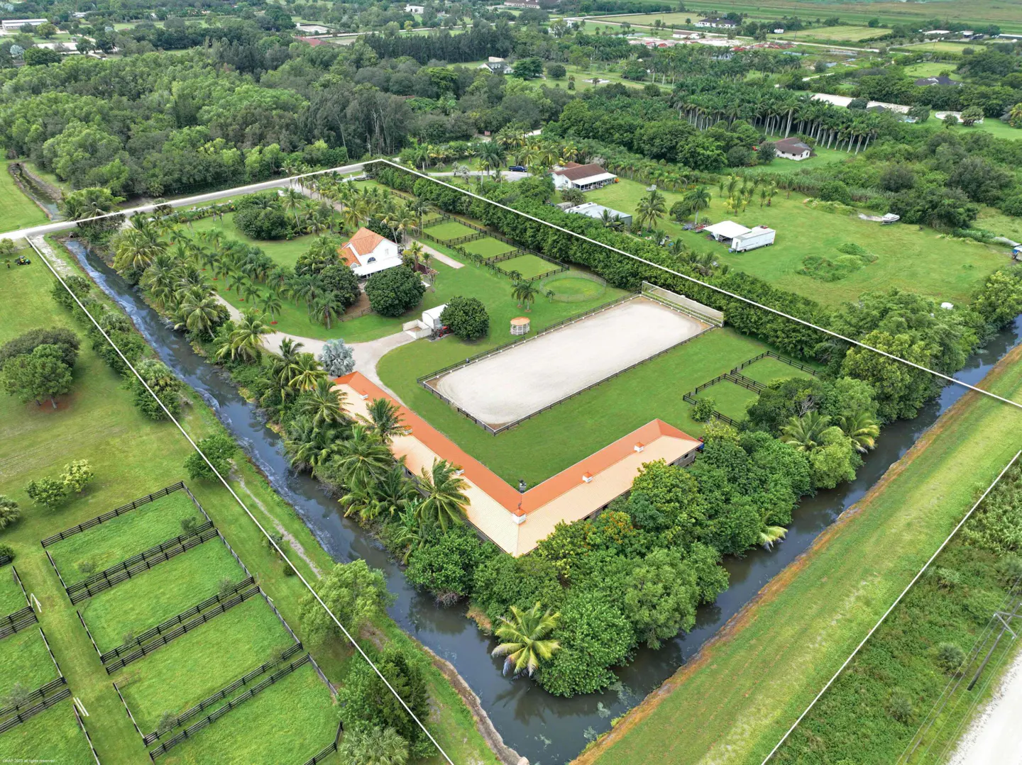 Aerial view of a horse farm with a white house, barn with orange roof, riding arena, and green pastures surrounded by trees and a canal.