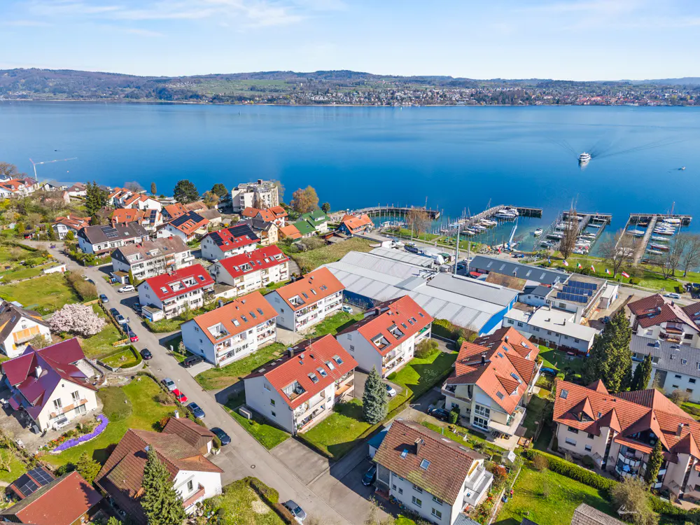 Aerial view of a lakeside town with red-roofed houses, a marina with boats, and a blue lake with distant hills.