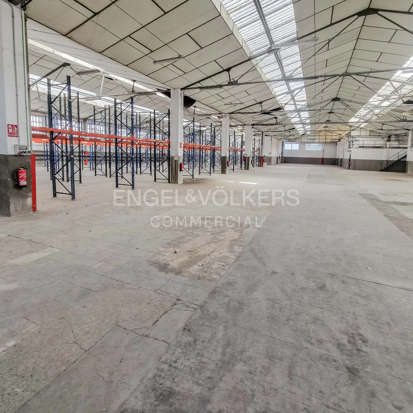 A large, empty warehouse with blue and orange shelving units. The floor is concrete and the ceiling is high with skylights.