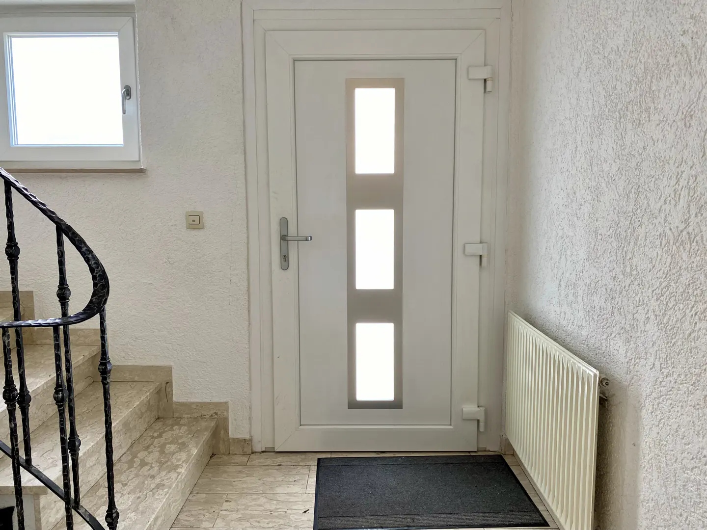 Interior view of a white front door with three vertical windows, a radiator, and a staircase with a black railing.