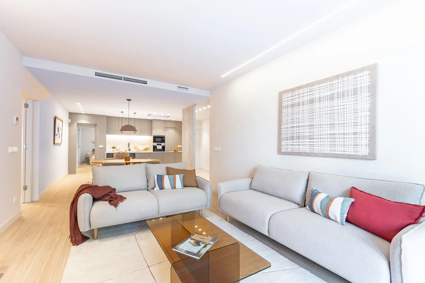 Modern living room with two light gray sofas, a glass coffee table, and abstract wall art, opening to a kitchen area.