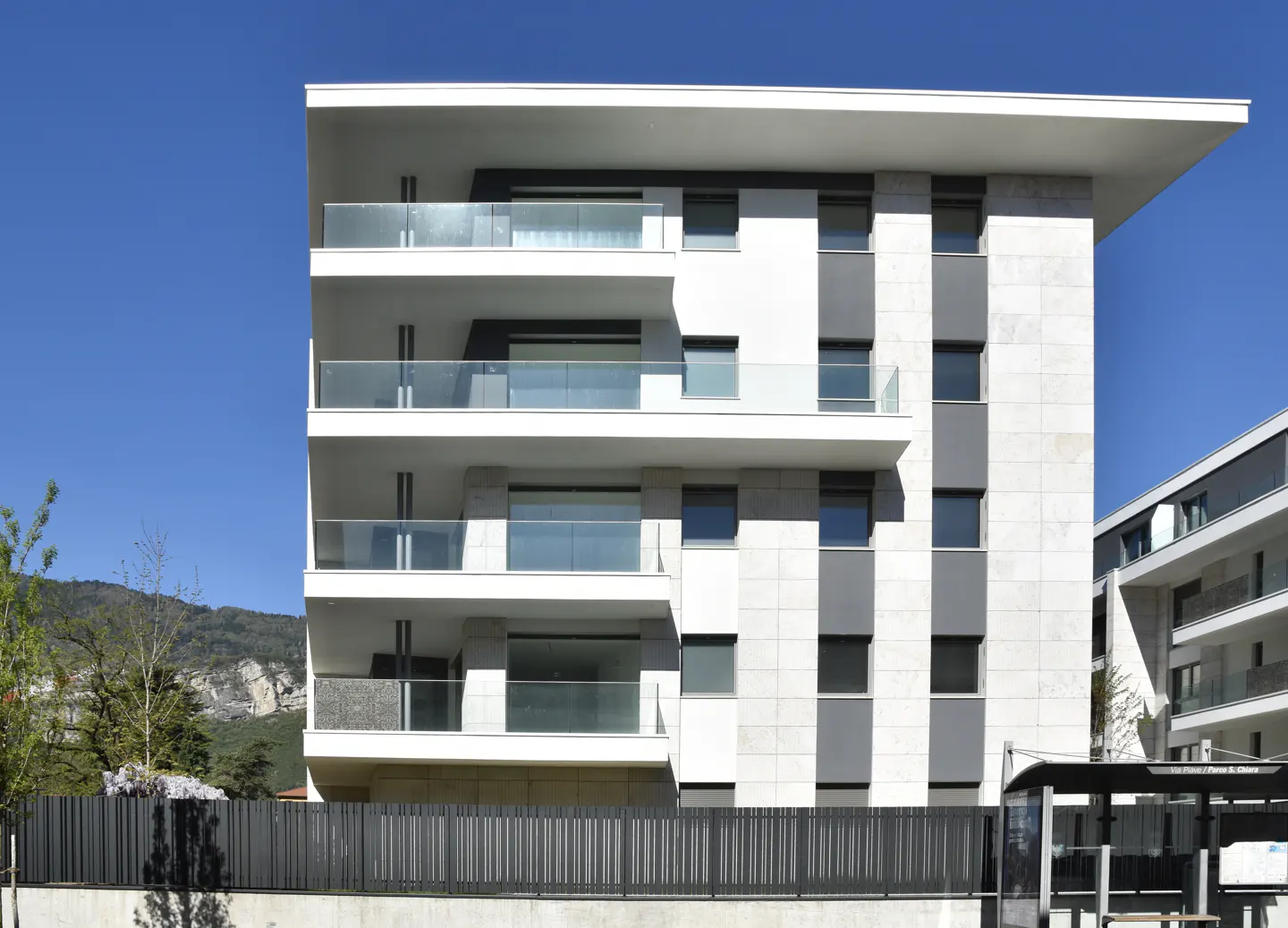 Modern white apartment building with glass balconies under a clear blue sky. A black fence and greenery are in the foreground.
