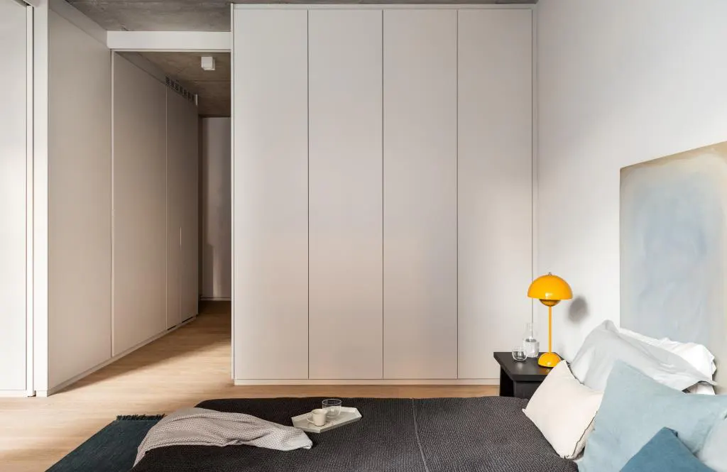 Bedroom with a gray bed, white pillows, and a yellow lamp on a black nightstand. White closets line the wall.
