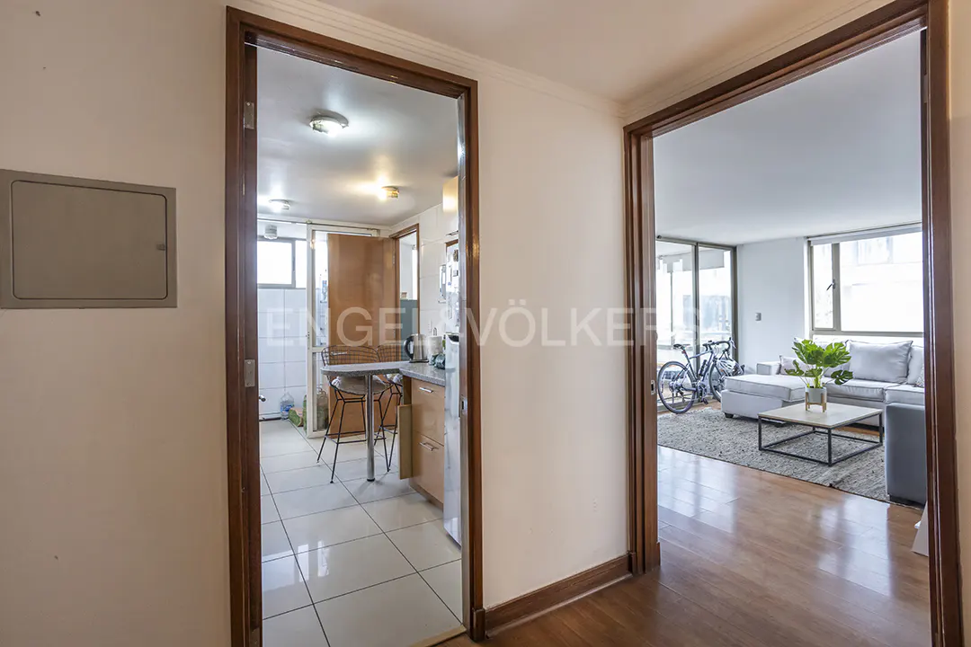A view through two doorways into a kitchen and living room. The kitchen has white tile and a small table. The living room has a couch, bike, and wood floors.
