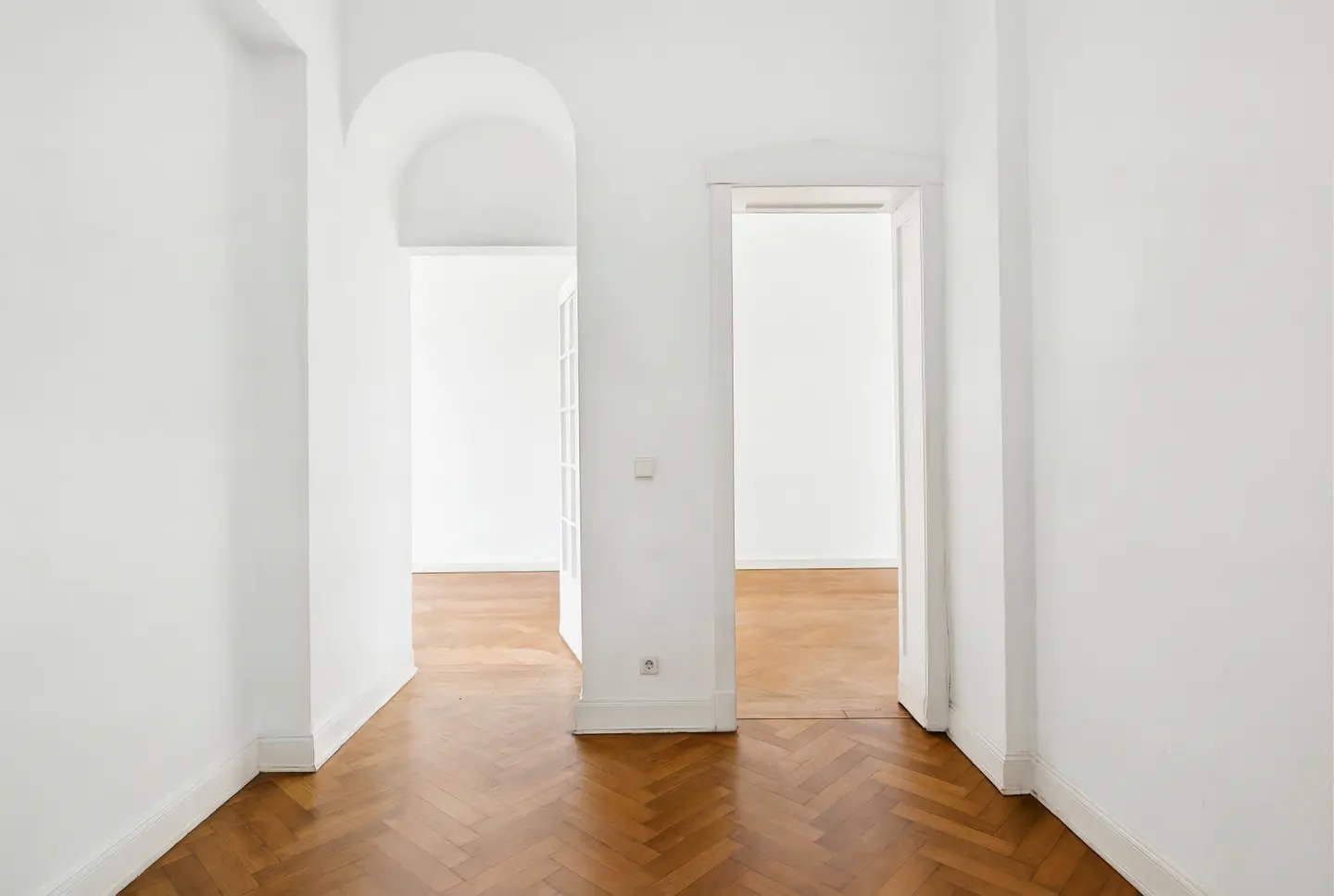 Interior view of a bright, empty room with white walls, herringbone wood floors, and two open doorways.