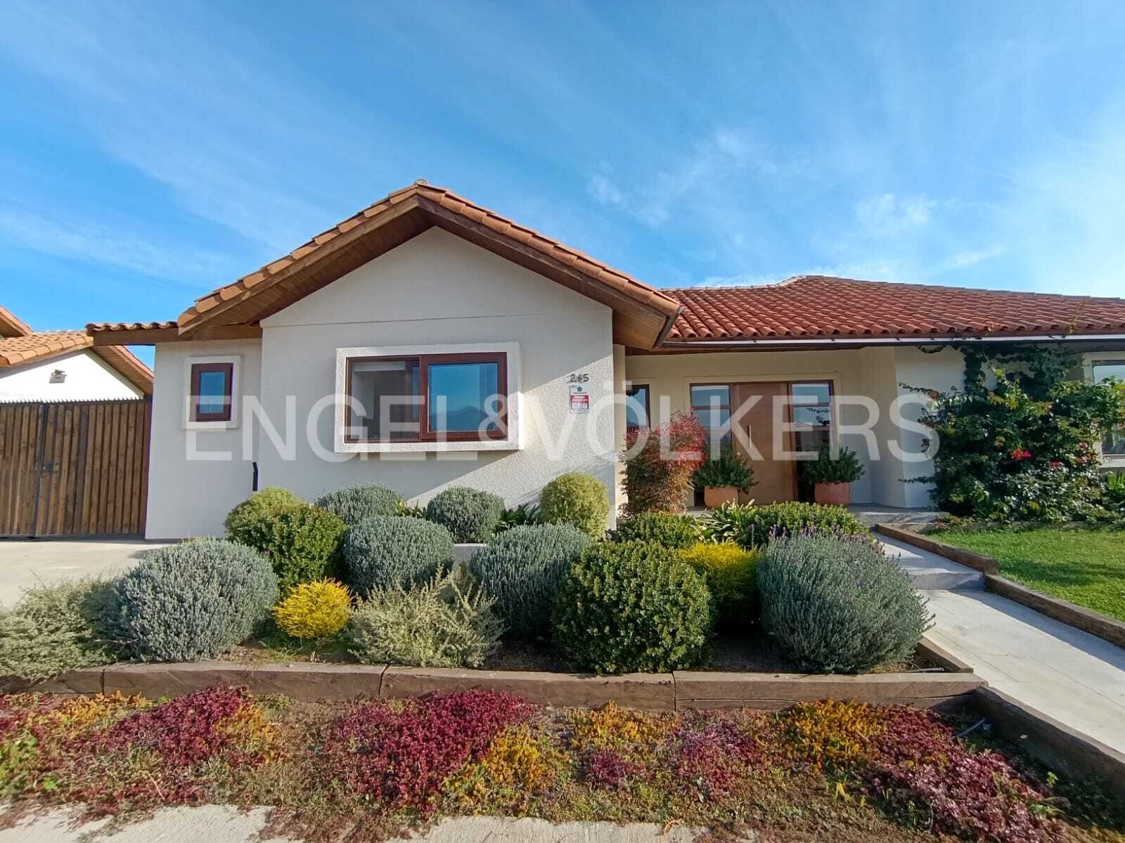 Single-story beige house with a terracotta tile roof, manicured round shrubs, and colorful ground cover under blue sky.