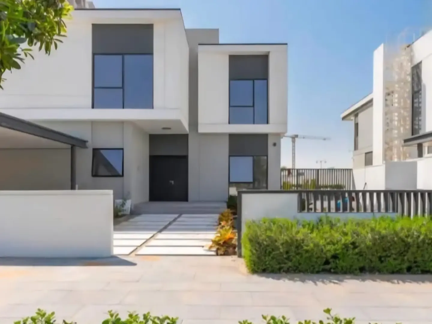 Modern two-story white house with black windows and door, green bushes, and a paved walkway on a sunny day.
