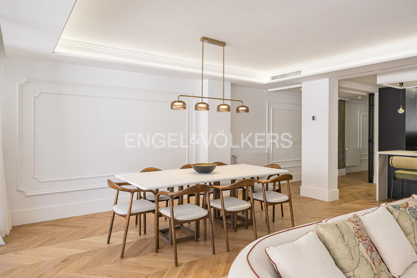 Bright dining room with white walls, herringbone wood floors, and a white table with eight chairs. A modern gold light fixture hangs above the table.