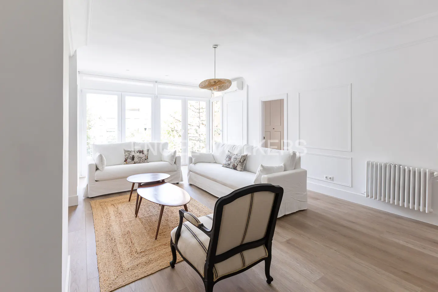 Bright living room with white sofas, wood floors, and a jute rug. A black-framed chair faces the seating area. Natural light streams through large windows.