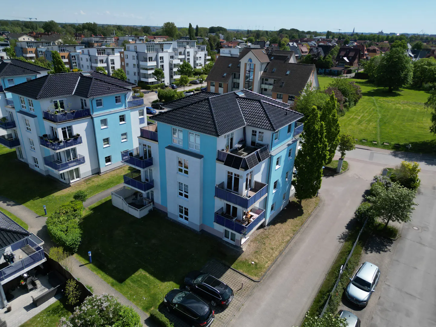 Aerial view of modern light blue and white apartment buildings with dark roofs, balconies, and surrounding green lawns.