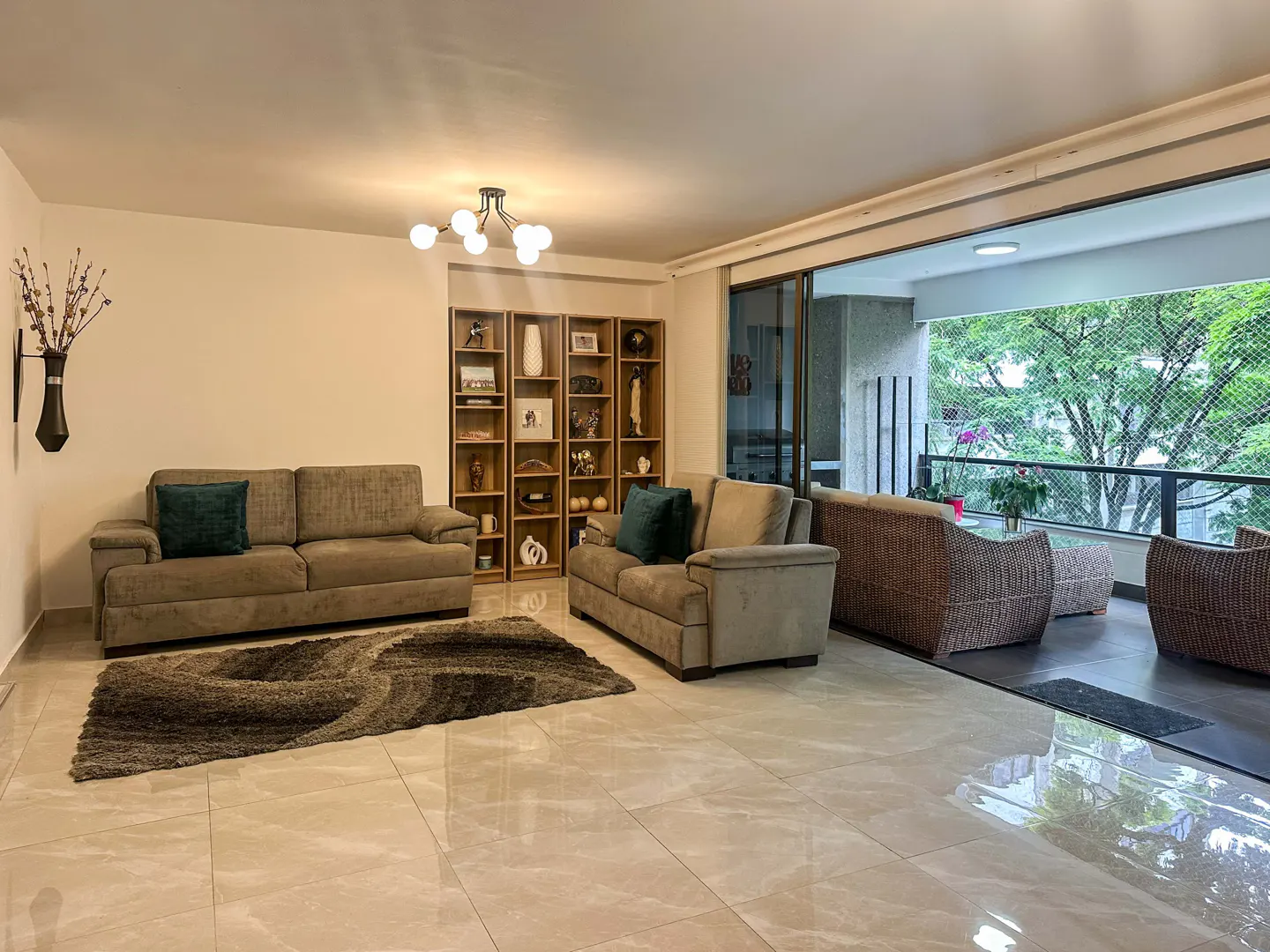Living room with beige sofas, a brown rug, and a wooden bookshelf. A balcony with wicker furniture overlooks green trees.