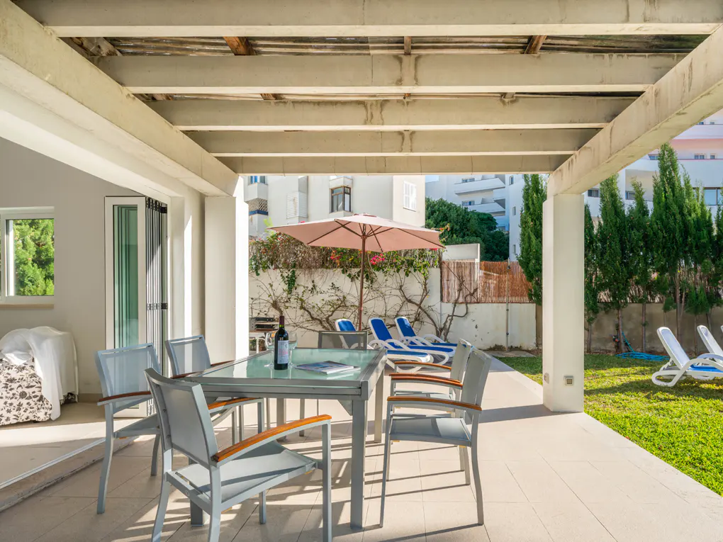 Outdoor patio with a glass table, chairs, wine bottle, and lounge chairs under an umbrella, leading to a green lawn.
