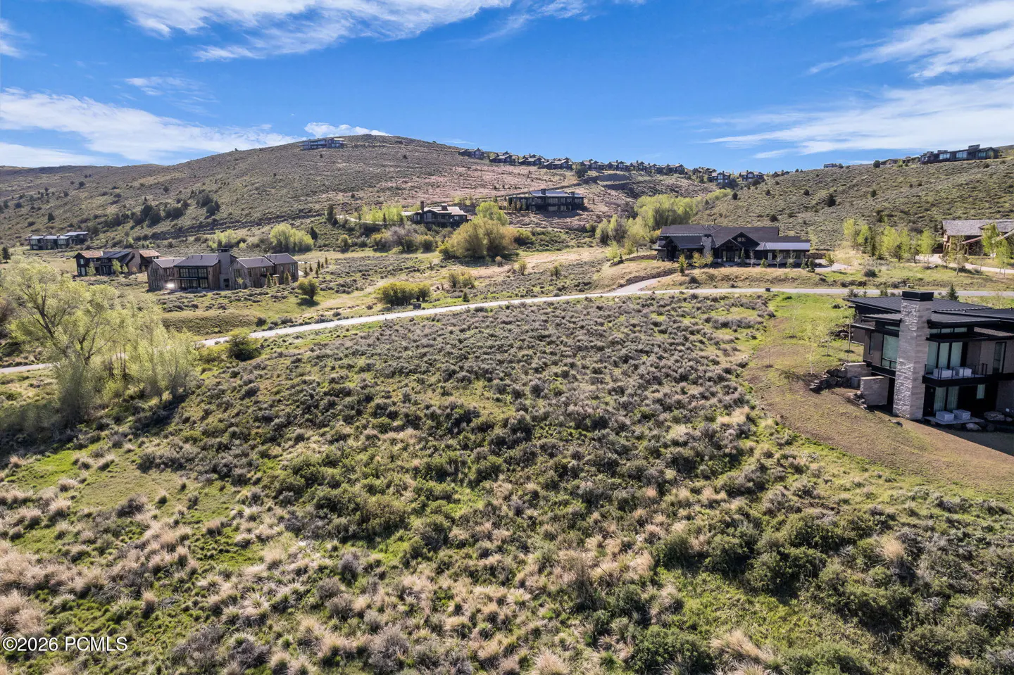 Hillside view of modern homes. Green and brown vegetation covers the foreground. A road winds through the landscape under a blue sky with clouds.