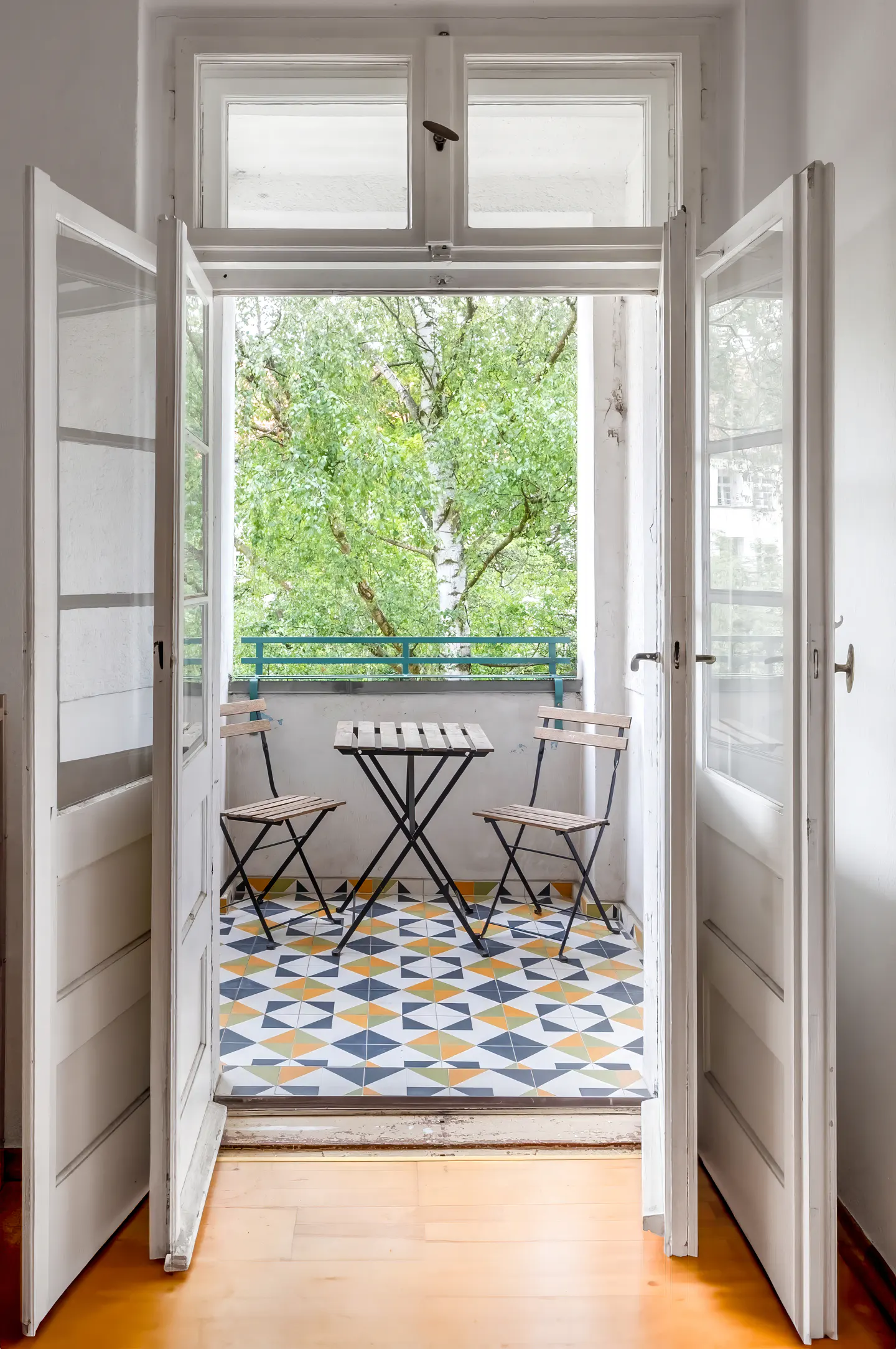 Open white doors reveal a balcony with a geometric tiled floor, a small table, two chairs, and lush green trees outside.
