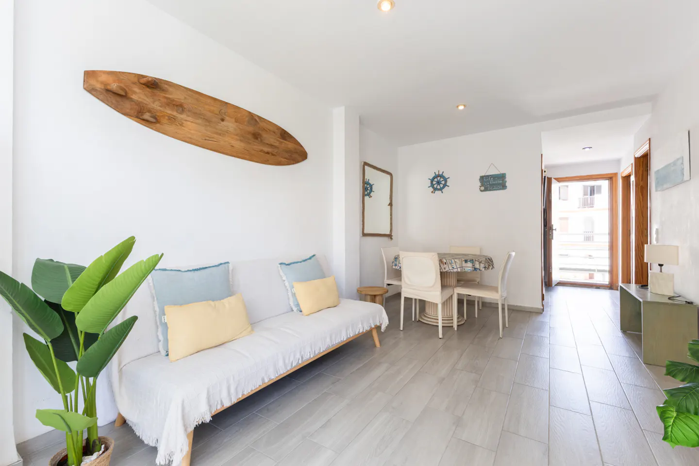 Bright living room with white walls, gray wood floors, and beach decor. A surfboard hangs above a white sofa with blue and yellow pillows.