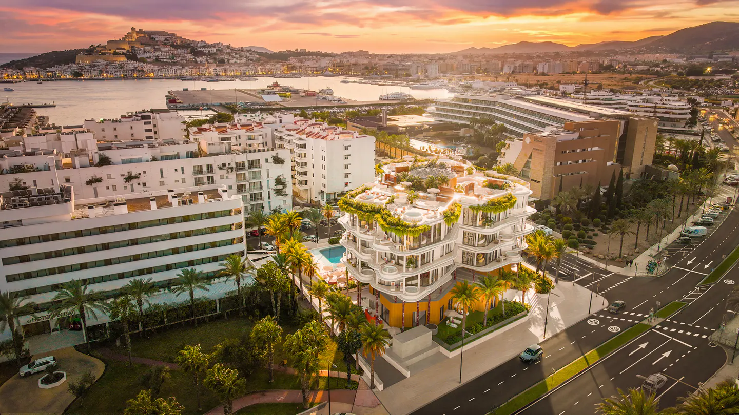 Aerial view of a modern white building with green plants, a pool, and palm trees, set against a sunset sky and coastal city backdrop.
